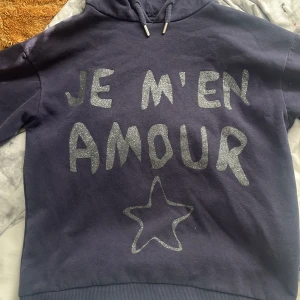 Mörkblå hoodie med glittertext - Säljer en mörkblå hoodie från ginatriot storlek 146/152 med huva och snörning. På framsidan står det 'JE M'EN AMOUR' i glittrig silvertext och en stjärna under. Tröjan har långa ärmar och ribbade muddar. Perfekt för dig som gillar coola statement-plagg med lite extra bling.