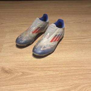 Adidas F50 fotbollsskor grå/blå/röd - Säljer ett par Adidas F50 fotbollsskor i grått med blå och röda detaljer. Skorna har en strömlinjeformad design utan snörning, med blå insida och klassiska Adidas-ränder på sidorna. Perfekta för dig som vill sticka ut på planen.