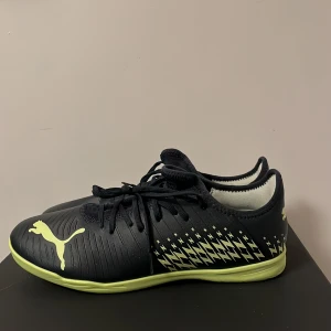 Svarta och gula Puma fotbollsskor - Snygga svarta fotbollsskor från Puma med coolt blixtmönster i gult på sidorna och gula detaljer på sulan. Skorna har snörning och är tillverkade i syntetmaterial, perfekta för inomhusspel. Puma-loggan syns tydligt både på ovansidan och sulan. Använda en gång eftersom att de var för stora.