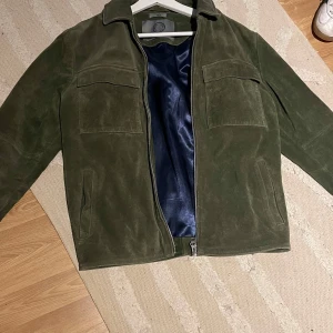 Human Scales. Olivgrön mockajacka  - Snygg olivgrön mockajacka från Human Scales i storlek S. Jackan har två stora bröstfickor med lock, dragkedja framtill och ett lyxigt mörkblått satinfoder. Perfekt för dig som gillar stilren och tidlös design. Jackan är helt oanvänd, säljes för att den inte passar.