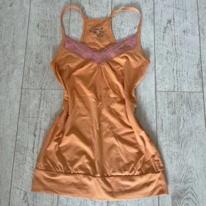 Persikofärgad linnetopp med spets - Snygg persikofärgad linnetopp från Multi Love med tunna axelband och v-ringning dekorerad med rosa spets. Toppen är figurnära och har bredare kant nedtill. Perfekt för en somrig look och superbekväm tack vare bomullsmaterialet med lite stretch.