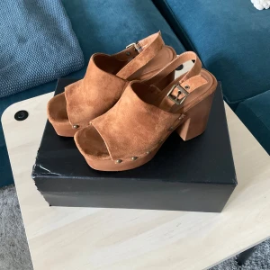 Bruna mockasandaler med klack och platå - Snygga sandaler i brun mocka med bred klack och platåsula. Skorna har öppen tå, justerbart spänne och coola nitar längs sidorna. Perfekta för dig som vill ha en retrovibe och sticka ut lite extra.