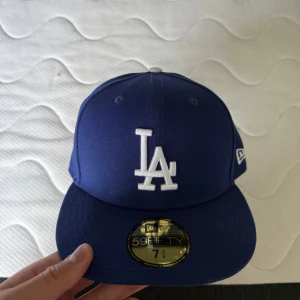 Blå LA Dodgers keps från New Era - Säljer en klassisk blå LA Dodgers 59FIFTY keps från New Era. Kepsen har broderad LA-logga framtill och MLB-logga bak. Den är i marinblå färg med böjd skärm och är tillverkad i polyester. Perfekt för dig som vill ha en ikonisk streetstyle-look.