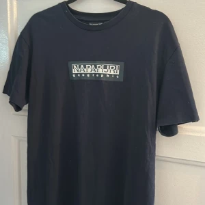 Svart Napapijri t-shirt med tryck - Svart t-shirt från Napapijri med klassiskt tryck på bröstet där det står 'Napapijri geographic'. Modellen har rund hals och korta ärmar. Materialet är mjuk bomull som känns skön mot huden. Perfekt för dig som gillar stilrena och enkla plagg med snygg logga. Nypris 399kr knappt använd 