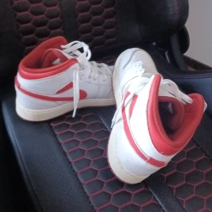 Nike Air Jordan 1 Mid vit/röd - Nike Air Jordan 1 Mid i vitt och rött med klassisk röd swoosh och detaljer. Skorna har snörning, perforerad tå och röd insida. Ovandel i skinn och syntet, platt sula och rund tå. Perfekt för dig som vill sticka ut med en ikonisk sneaker.