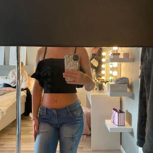 Svart croppad volangtop med spets - Supersöt svart croppad topp med tunna axelband och volangdetaljer. Toppen har broderade prickar i tyget och är tillverkad i bomull. Spetskänsla och dragkedja bak gör den extra snygg till jeans eller kjol.