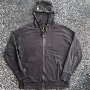 C.P. Company zip hoodie - Svart hoodiejacka från C.P. Company med ikoniska goggles i huvan. Jackan har dragkedja framtill, två fickor med dragkedja och en relaxed passform. Tillverkad i mjuk bomull, perfekt för en streetwear-look. Storlek L, har två hål på ryggen annars bra skick 7/10, pris går att diskutera✅