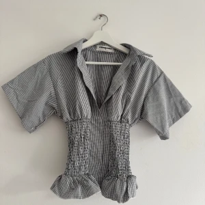 Rutig figursydd skjorta - Superfin svartvit rutig skjorta från Missguided i storlek XS. Skjortan har korta ärmar, bred krage, smockad midja och volangkant nedtill. Perfekt för dig som gillar en trendig och unik look med markerad midja. Endast testad! 