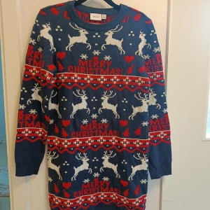 Blå julklänning med renar från Vila - Rolig blå stickad julklänning från Vila med mönster av vita renar, snöflingor, hjärtan och texten 'Merry Christmas!' i rött. Klänningen har långa ärmar och ribbad kant vid hals och ärmslut. Perfekt för julens alla tillfällen!