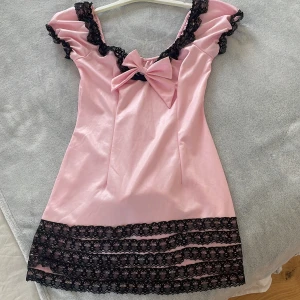 Rosa kort klänning med spets och rosett - Supersöt rosa kort klänning med svarta spetsdetaljer vid axlar och nederkant. Stor rosett framtill och baktill som gör den jättegullig. Lappen är bortklippt så vet inte märke eller exakt storlek. Men skulle säga att den passar S och M då tyget har stretch.