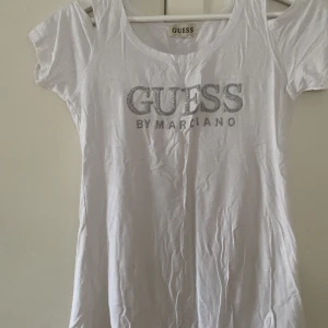 Vit t-shirt från Guess - Säljer en vit t-shirt från Guess med glittrig logga framtill. T-shirten har korta ärmar och rund halsringning. Perfekt för en avslappnad stil.
