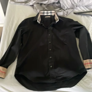 Svart skjorta med rutiga detaljer från Burberry - Snygg svart skjorta från Burberry med klassiska rutiga detaljer på krage och ärmslut. Skjortan har knappar framtill och en diskret broderad logga. Perfekt för en stilren look.