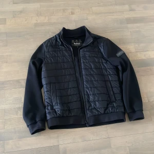 Svart quiltad jacka från Barbour International - Snygg svart jacka från Barbour International med quiltade detaljer på framsidan och släta ärmar. Jackan har en dragkedja framtill och en stilren design som passar perfekt för höst och vår. Märkeslogga på ärmen ger en extra touch av stil.