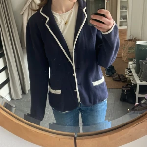 Mörkblå kavaj från Lexington - Säljer en stilren mörkblå kavaj från Lexington med vita detaljer. Kavajen har knappar framtill och vita kanter vid krage och fickor. Perfekt för en elegant look.