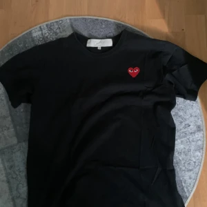 Svart t-shirt från Comme des Garçons - Säljer en svart t-shirt från Comme des Garçons med det ikoniska röda hjärtat med ögon broderat på bröstet. T-shirten är tillverkad i 100% bomull och har en klassisk passform. Perfekt för en stilren look.