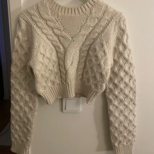 Beige stickad tröja - Säljer en mysig beige stickad tröja med flätmönster och ribbade muddar. Tröjan är croppad och har långa ärmar, perfekt för kyliga dagar.