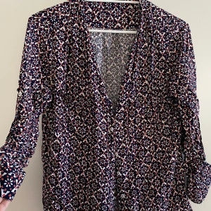 Mönstrad blus med knappar - Snygg blus med ett detaljerat geometriskt mönster i blått, vitt och rött. Blusen har långa ärmar och knappar framtill, perfekt för en stilren look.