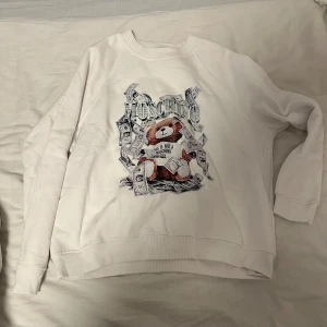 Vit sweatshirt från Moschino med nallemotiv - Säljer en vit sweatshirt från Moschino med ett coolt nallemotiv omgivet av sedlar. Tröjan har långa ärmar och en rund halsringning. Perfekt för en avslappnad stil med en touch av lyx. Pris sänks vid snabbaffär.