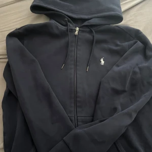 Mörkblå huvtröja med dragkedja från Ralph Lauren - Säljer en stilren mörkblå huvtröja från Ralph Lauren med dragkedja och broderad logga på bröstet. Tröjan har långa ärmar och en praktisk huva med snörning. Perfekt för en avslappnad look.