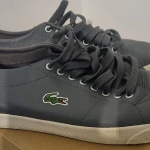 Grå sneakers från Lacoste - Snygga grå sneakers från Lacoste med klassisk krokodillogga på sidan. Skorna har snörning och en vit sula som ger en stilren look. Perfekta för en avslappnad stil. Mycket bra skick 