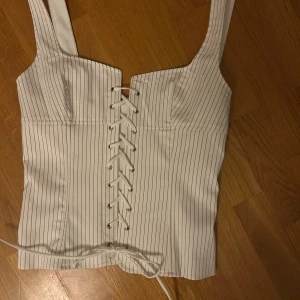 Vit randig korsettopp - Snygg vit korsettopp med smala svarta ränder och snörning framtill. Toppen har breda axelband och en dragkedja bak för enkel påklädning. Perfekt för en stilren look.