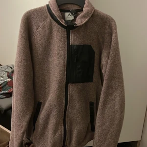 Brun fleecejacka från Mountain Warehouse - Mysig brun fleecejacka från Mountain Warehouse med svarta detaljer. Jackan har en dragkedja framtill och praktiska fickor. Perfekt för kyliga dagar.