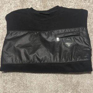 Svart tröja med läderdetaljer från Prada - Snygg svart tröja från Prada med läderdetaljer över bröstet och en liten dragkedja. Tröjan har långa ärmar och en rund halsringning. Perfekt för en stilren look.