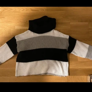 Randig stickad polotröja - Säljer en mysig stickad polotröja med breda ränder i svart, grått och vitt. Tröjan har en hög polokrage och långa ärmar, perfekt för kyliga dagar. Den ribbade strukturen ger en skön passform.