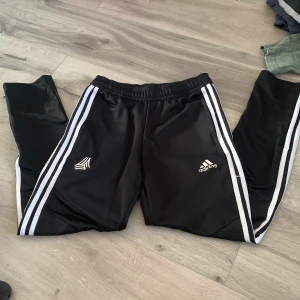 Svarta träningsbyxor från Adidas - Säljer ett par svarta träningsbyxor från Adidas med de klassiska tre vita ränderna längs sidorna. Byxorna har en elastisk midja och är perfekta för sport och träning. Adidas-loggan är tryckt på framsidan. * Barnsize 