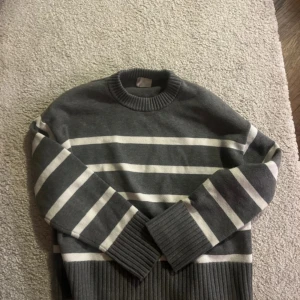 Grå och vit randig tröja - Säljer en stilren grå tröja med vita ränder. Tröjan har långa ärmar och ribbade muddar vid ärmslut och nederkant. Perfekt för en avslappnad look.