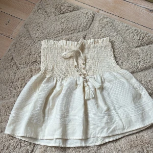 Beige kjol från Zara - Charmig beige kjol från Zara med smockdetaljer i midjan och dekorativa knappar framtill. Kjolen har en kort längd och är tillverkad i ett lätt och luftigt material, perfekt för varmare dagar. Den har även en fin knytning med tofsar som ger en bohemisk touch.