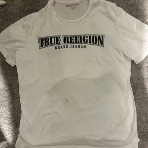 Vit t-shirt från True Religion - Säljer en vit t-shirt från True Religion med svart text på framsidan och ett stort svart tryck på baksidan. Perfekt för en avslappnad stil. T-shirten har korta ärmar och en rund halsringning. Storlek medium. Bra skick förutom en liten fläck på framsidan som inte syns så mycket. Annars bra skick. Köptes i NK Göteborg. Kvitto och tags finns kvar.