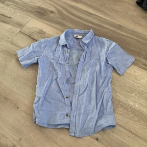 Blå kortärmad skjorta från Pomp de Lux - Säljer en ljusblå kortärmad skjorta från Pomp de Lux. Skjortan har en klassisk krage och knappar framtill. Perfekt för varma dagar och ger en stilren look. Passar bra till både jeans och shorts.*Barnsize