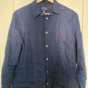 Blå linneskjorta från Ralph Lauren - Säljer en stilren blå linneskjorta från Ralph Lauren med klassisk krage och knappar framtill. Skjortan har en broderad logotyp i rött på bröstet och långa ärmar. Perfekt för en avslappnad men ändå elegant look. På lappen står det xl men själv tycker jag att den sitter som en s. Bra skick 