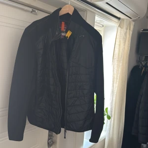 Svart jacka från Parajumpers - Säljer en svart jacka från Parajumpers med quiltad design och dragkedja. Jackan har en gul detalj vid kragen och ett märke på ärmen. Perfekt för kyligare dagar.