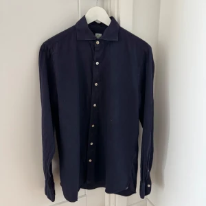 Mörkblå skjorta från Finamore Napoli - Säljer en stilren mörkblå skjorta från Finamore Napoli med klassisk krage och knappar framtill. Skjortan har långa ärmar och en diskret broderad detalj vid ärmslutet. Perfekt för en elegant look.