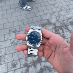 Tissot herrklocka med blå urtavla - Snygg Tissot herrklocka med en stilren blå urtavla och datumvisning. Klockan har ett robust armband i silverfärgat stål och en transparent baksida som visar urverket. Perfekt för den som uppskattar både design och funktion.