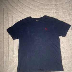 Mörkblå t-shirt från Polo Ralph Lauren - Pris diskurterbart! Detta är en klassisk mörkblå t-shirt från Polo Ralph Lauren med det ikoniska röda logotypbroderiet på bröstet. T-shirten är kortärmad och tillverkad i mjuk bomull för en bekväm passform. Perfekt för en avslappnad stil.