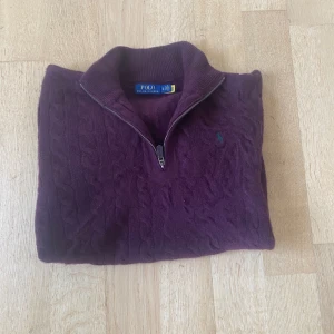 Stickad Polo Ralph lauren tröja - Säljer en stilren vinröd stickad tröja från Ralph Lauren med dragkedja vid halsen. Tröjan har ett diskret märke på bröstet och är perfekt för kyligare dagar. Den har långa ärmar och ribbade muddar för en bekväm passform.