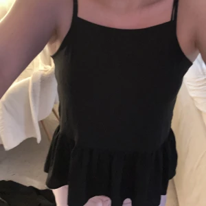 Svart ärmlös topp - Säljer en stilren svart ärmlös topp med smala axelband och volangdetalj vid nederkanten. Perfekt för en chic och enkel look. Passar bra till både jeans och kjol.