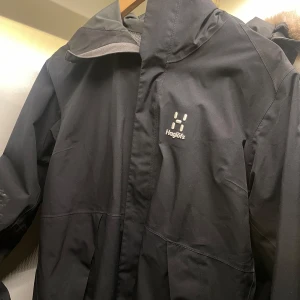 Svart vindjacka från Haglöfs - Säljer en svart vindjacka från Haglöfs med dragkedja och hög krage. Jackan har en stilren design med Haglöfs-logga på bröstet och är tillverkad i ett vattentätt material. Perfekt för blåsiga dagar och svala sommarkvällar. 