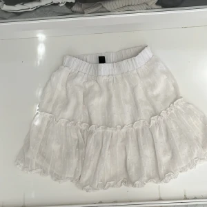 Vit kjol med volang - Säljer en söt vit kjol med volangdetaljer. Kjolen har ett elastiskt midjeband och ett diskret prickigt mönster som ger en fin textur. Perfekt för varma dagar och kan enkelt matchas med olika toppar. 🌸
