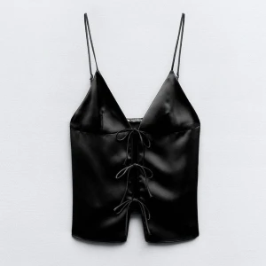 Svart satinlinne med snörning - Snyggt svart linne i satin med smala axelband och dekorativ snörning framtill. Perfekt för en stilren och elegant look. Liten slitning vid ena snöret