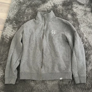 Grå zip-up hoodie från Les Duex - Säljer en stilren grå zip-up hoodie från Les Duex. Tröjan har en dragkedja framtill och en liten logga på bröstet. Perfekt för en avslappnad stil.