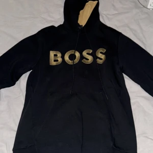 Svart hoodie från BOSS - Säljer en stilren svart hoodie från BOSS med guldigt broderat märke på bröstet. Hoodien har en justerbar huva med beige insida och en praktisk magficka framtill. Perfekt för en avslappnad look. Xs-s