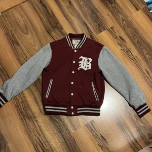 Vinröd och grå Collagejacka   - Unik vinröd  Collagejacka med grå ärmar från Seven Teen. Jackan har ett stort 'B' broderat på bröstet och stängs med knappar. Storlek står M men sitter som S 