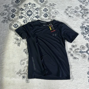 Svart t-shirt från Under Armour - Säljer en svart t-shirt från Under Armour, perfekt för träning. Den har en normal passform och är kortärmad med en diskret logga på bröstet. Materialet är syntetiskt och andas bra, vilket gör den idealisk för intensiva aktiviteter.