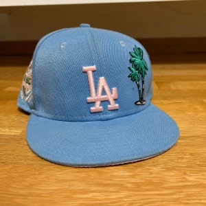 Blå keps med broderad palm och LA-logga - Snygg blå keps från New Era med broderad LA-logga i rosa och en palm i grönt. Kepsen har även ett jubileumsmärke på sidan och en liten logga bak. Perfekt för att ge din stil en cool touch! Knappt använd. 