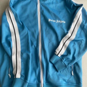 Blå träningsjacka från Palm Angels - Snygg blå träningsjacka från Palm Angels med vita ränder längs ärmarna. Jackan har dragkedja framtill och två fickor med dragkedjor. 