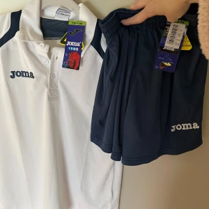 Vit kortärmad piketröja och blå shorts  - Säljer en vit kortärmad piketröja från Joma med mörkblå detaljer på axlarna. Piketröja har en klassisk krage med knappar och är gjord i ett lätt och bekvämt material. Och shorts i blå färg. Storlek passar barn 10-14 år